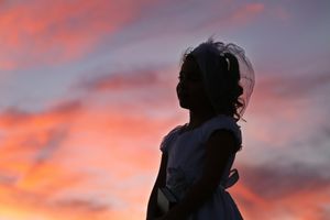 Silhouette of a girl in a white dress and veil against a vivid pink and orange sunset sky.