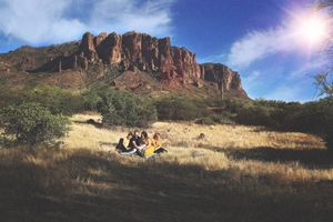 Group of people relaxes on a grassy hillside in the sun with towering red rock cliffs and a bright blue sky behind them.