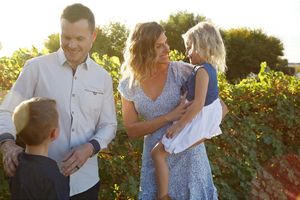 Smiling family of four stands in front of green grapevines as a mother holds her young daughter and a dad greets his son.
