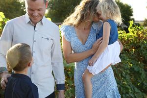 Smiling family of four embraces outdoors among green foliage in warm golden light.