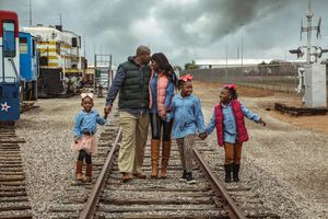 Parents and three daughters walk hand in hand along railroad tracks past train cars on a cloudy day.