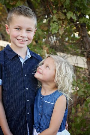 Smiling boy in a navy polka dot shirt stands beside his younger blonde sister in a denim top, who gazes up at him outdoors.