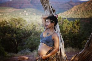 Pregnant woman with a headband stands beside a weathered tree trunk with red rock hills in the distance.