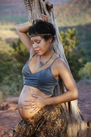 Pregnant woman in a gray top and patterned pants poses with a hand on her belly beside a rustic tree trunk.