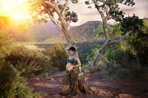 Pregnant woman in a boho outfit stands beside a twisted tree at sunset overlooking red rock hills in Sedona.