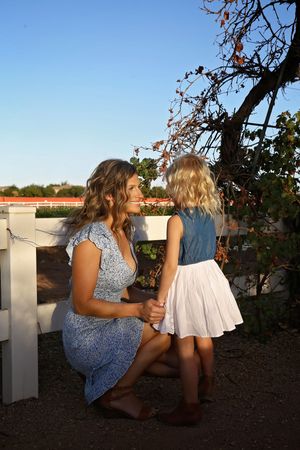 Mother in a blue floral dress kneels to hold hands with her young daughter beside a white fence under a clear blue sky.
