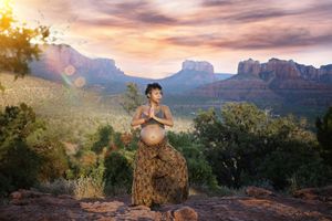 Pregnant woman in patterned wide-leg pants holds a prayer pose on a rocky ledge with red rock buttes at sunset.
