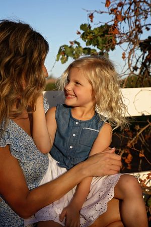 A smiling blonde girl in a denim top and white skirt sits on a fence beside her mother in warm evening light.