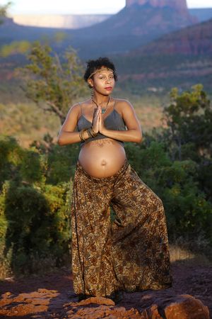 Expectant mother in a gray top and printed pants stands in a yoga pose with her hands at heart center in the desert.