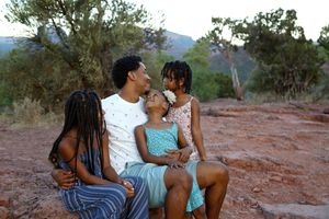 Smiling father sits on red rocks hugging three daughters in a warm outdoor family portrait at sunset.