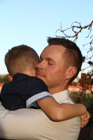 A smiling man with closed eyes tenderly hugs a young boy in a navy shirt outdoors at golden hour.