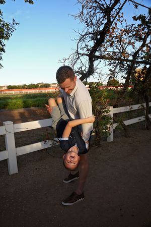 A man in a gray shirt playfully holds a laughing boy upside down on a dirt path beside a white ranch fence.