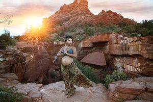 Pregnant woman in a blue top and flowing patterned skirt poses on red rocks as the sun sets behind a rugged peak.
