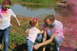 Two children and an adult in sunglasses play on a grassy bank near a pond amid pink powder during a color festival.