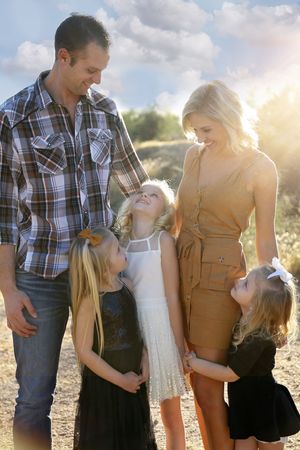 Family of five laughs together outdoors in golden sunlight, with parents hugging three young daughters in dresses.