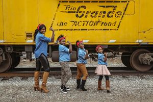 Mother and three daughters in denim shirts and red bandanas flex their arms in front of a yellow Rio Grande train car.