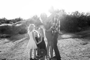 Black and white backlit portrait of parents embracing as three young daughters huddle close in a sunlit desert field.