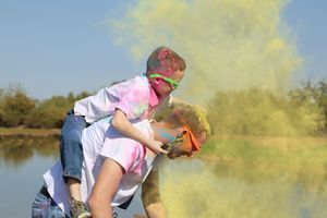 Two children in white shirts with pink color powder on their faces and clothes play piggyback near a lake as yellow powder clouds the air.