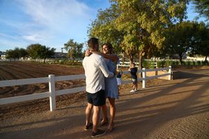 A couple embraces on a sunny dirt path beside a white wooden fence and a plowed field with trees in the background.