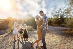 A couple embraces in a warm dance on a sunlit dirt path while three young girls in dresses hold hands and twirl nearby.