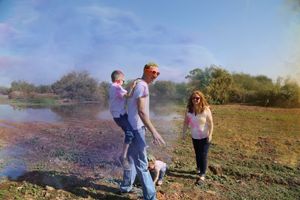 A smiling family walks along a grassy lakeshore, with a father carrying a child on his back and a mother beside them, in colorful powder.