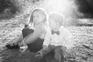 A black and white photo of a young girl and a boy in a bow tie sitting back to back on the ground in soft backlight.
