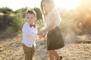 A little boy in a bow tie and suspenders holds hands with an older girl in a skirt as golden sunlight glows over a desert field.