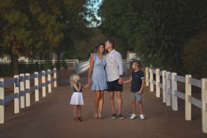 A family of four in coordinated outfits walks together along a white-fenced country path beneath tall green trees.