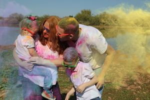A family of four in white shirts shares a happy hug, laughing with color powder on their clothes as yellow smoke drifts behind them.