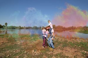 A family stands by a lake as colorful pink, blue and orange powder clouds fill the sky, with a child riding on a parent's shoulders.