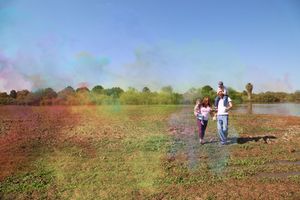A small family walks across a wide open field near a lake under a hazy blue sky tinted with soft clouds of colorful powder.
