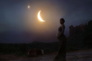 Silhouette of a pregnant woman in a long dress standing on a desert hillside beneath a glowing crescent moon at dusk.