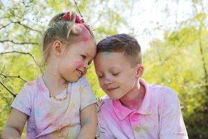 Young girl and boy with colorful powder paint on their faces and shirts sit close together, smiling in a sunny green park.