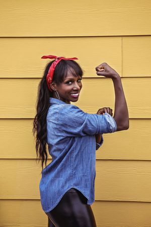 Smiling woman in a denim shirt and red bandana flexes her bicep in a classic Rosie the Riveter pose against a yellow wall.