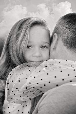 Black and white portrait of a girl in a polka dot blouse resting her chin on a man's shoulder as they hug under a cloudy sky.