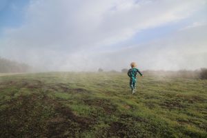 Small child in green pajamas runs through a grassy field covered in morning mist under a pale blue sky.