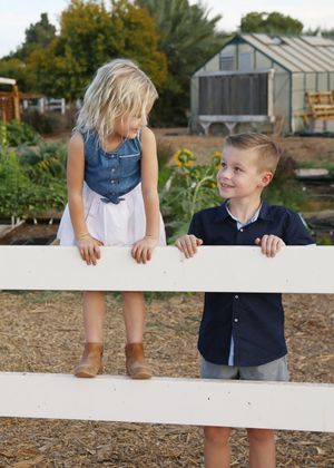 Blonde girl in a denim top stands on a white fence rail smiling at a boy in a navy shirt, with a garden and greenhouse behind.