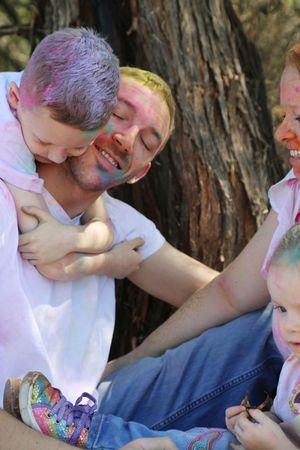 Smiling father with colorful powder paint on his face hugs a young boy with purple-dusted hair beside a tree with his family.