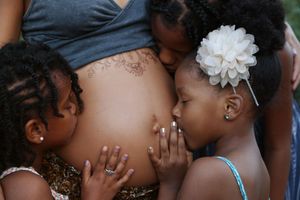 Two young girls with braided hair, one wearing a white flower, kiss and touch the bare pregnant belly of their mother.