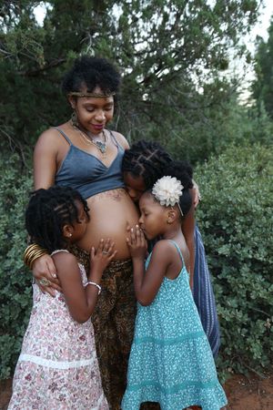 Pregnant mother in a headband and necklace stands in a green garden as three daughters in sundresses kiss and hug her belly.