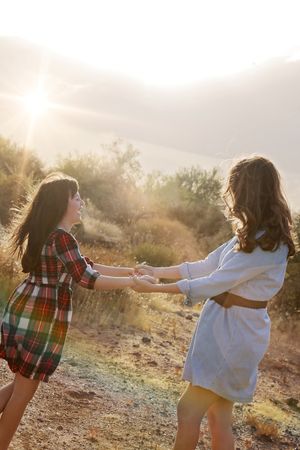 Two girls hold hands and spin joyfully in a sunlit desert landscape, one in a plaid dress and one in a blue dress.