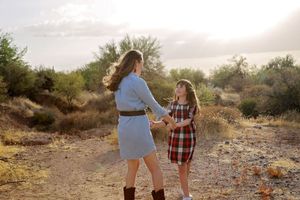 Mother and daughter hold hands and smile at each other on a sunny desert trail lined with green shrubs and trees.