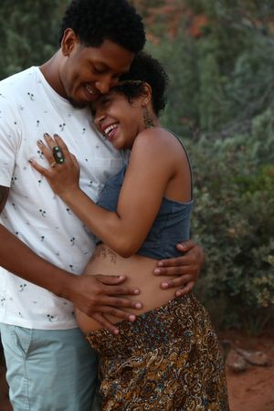 A smiling couple embraces closely outdoors, the man holding the woman's waist as she rests her head on his chest among green trees.