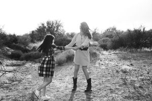 Two girls hold hands and spin in a joyful circle on a sunlit desert path in this black and white outdoor family photo.