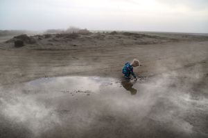 A small child in a blue jacket crouches beside a muddy puddle on a wide, misty beach under an overcast sky.