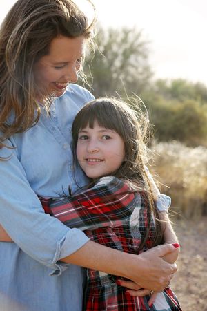 Smiling girl in a red plaid shirt is hugged by her laughing mother in warm golden light outdoors.