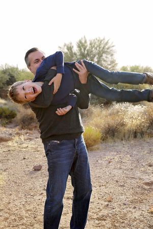 Playful father holds his laughing young son upside down in a sunny desert setting with dry grass and trees.