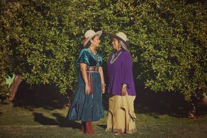 Two smiling women in wide-brimmed hats, teal and purple traditional outfits, and silver jewelry stand together before lush green trees.