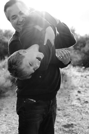 Black and white photo of a smiling man playfully holding a laughing boy upside down outdoors in soft backlight.