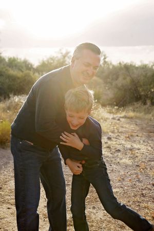 A smiling man in a dark sweater hugs and tickles a laughing boy on a desert trail in golden sunlight.
